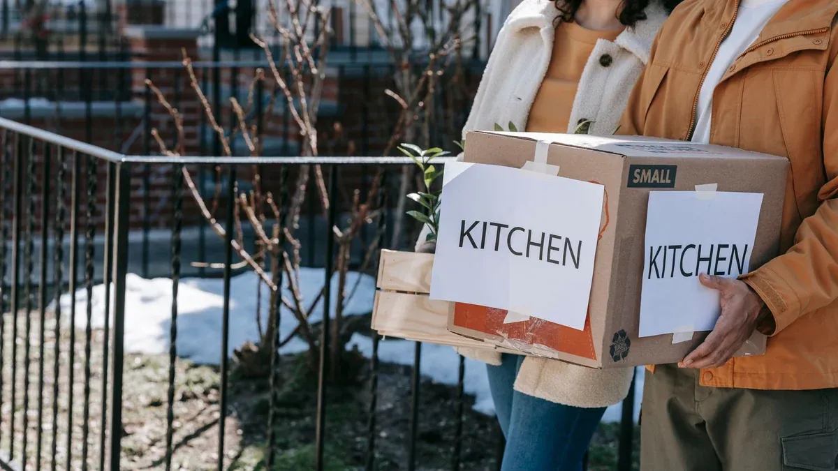 A couple carrying labeled kitchen boxes while moving into a new home outdoors.