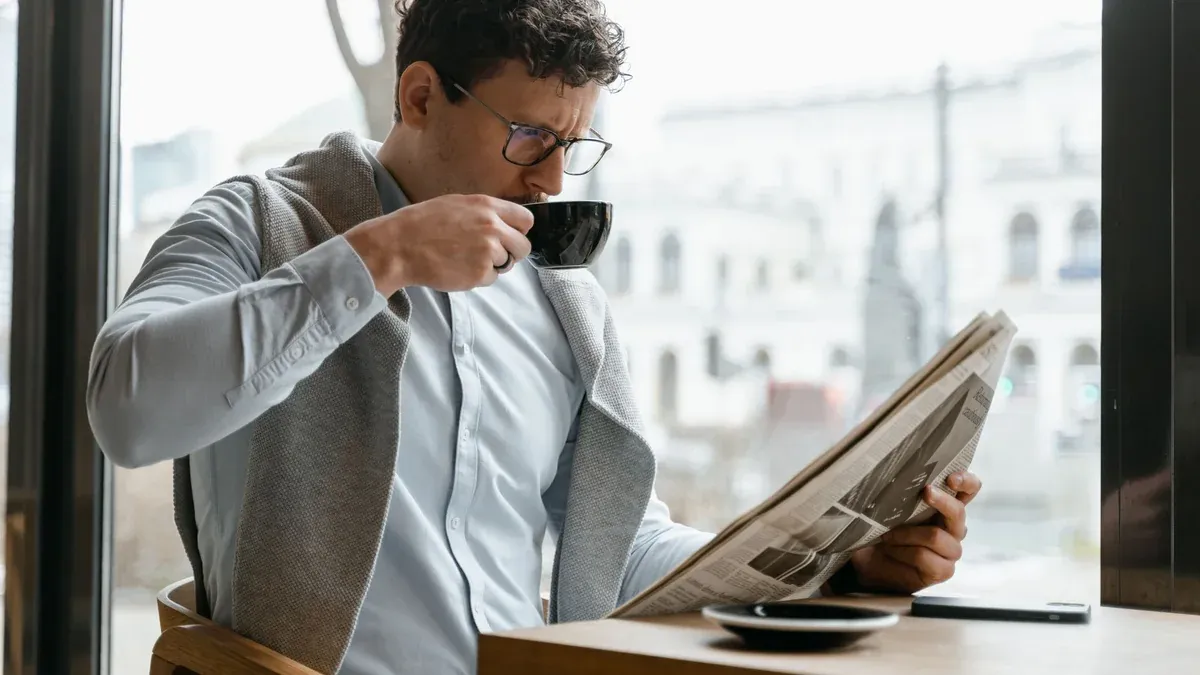 Adult man wearing eyeglasses sipping coffee and reading a newspaper in a cozy cafe setting.