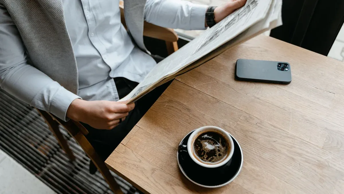 An adult male enjoying his coffee while reading a newspaper at a cafe with his smartphone nearby.