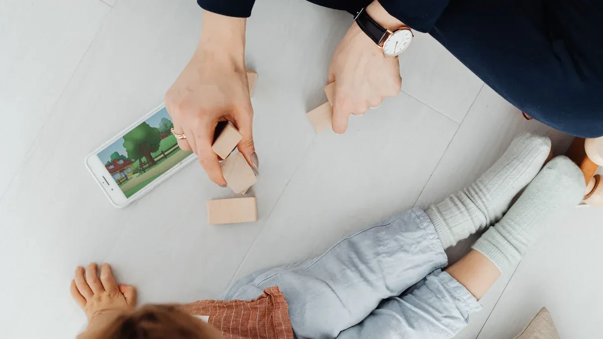 A mother and child playing with wooden blocks and a smartphone on the floor.