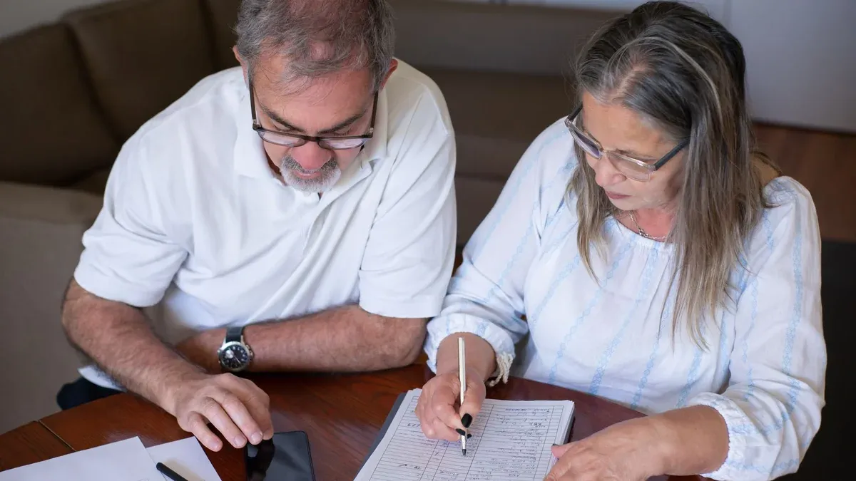 Elderly couple reviewing documents, using smartphone for online banking at home.