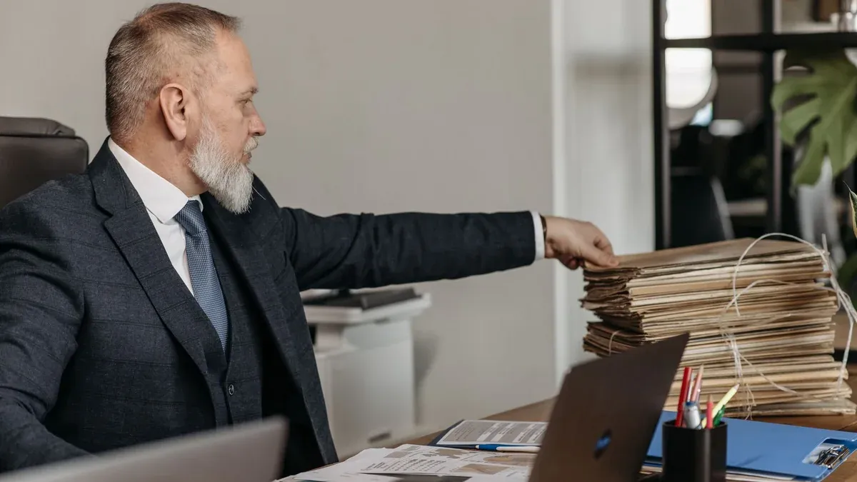 Mature businessman reaching for a stack of files on his desk in a modern office environment.
