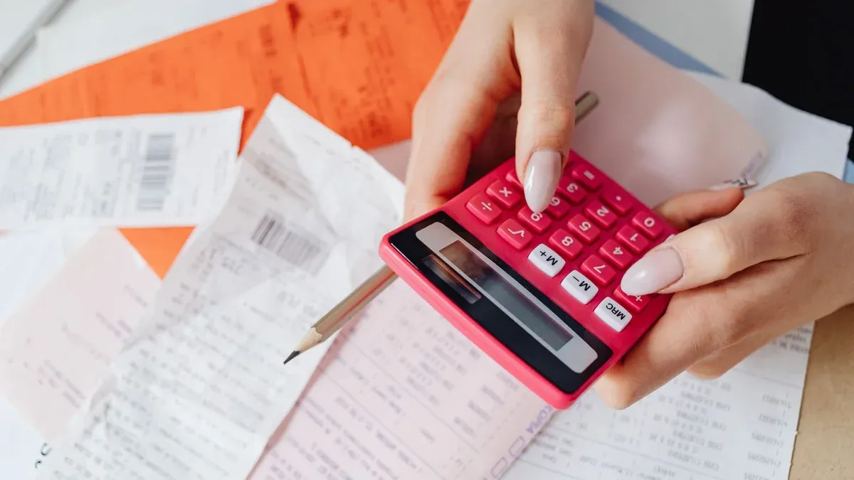Hands using a pink calculator to manage expenses amidst various receipts and documents.