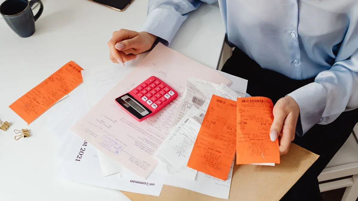 A woman reviews receipts and calculates expenses at a desk with a pink calculator.