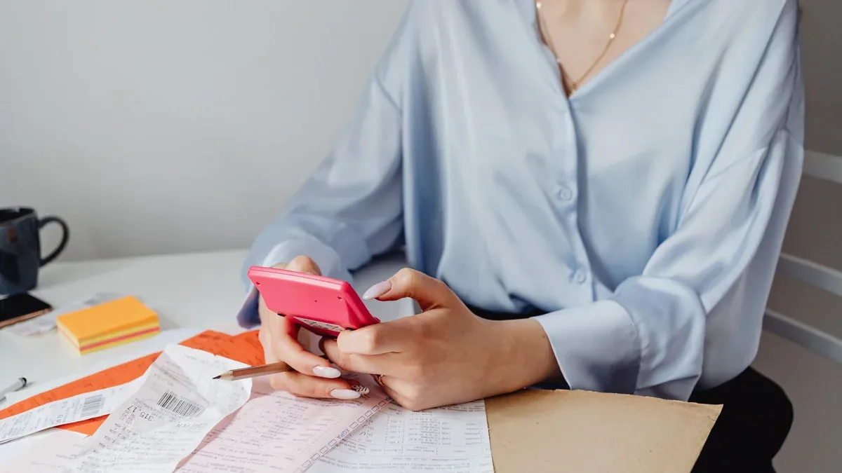 A woman using a pink calculator surrounded by bills and receipts at a desk.