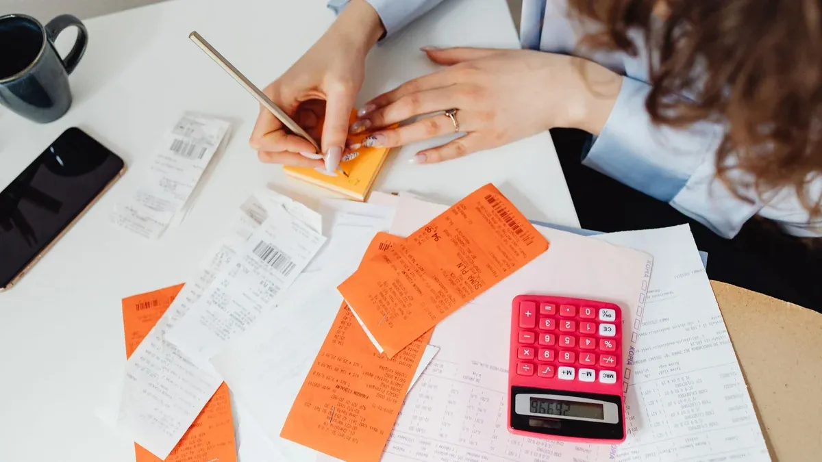 A woman writes down financial details using a calculator, receipts, and documents.