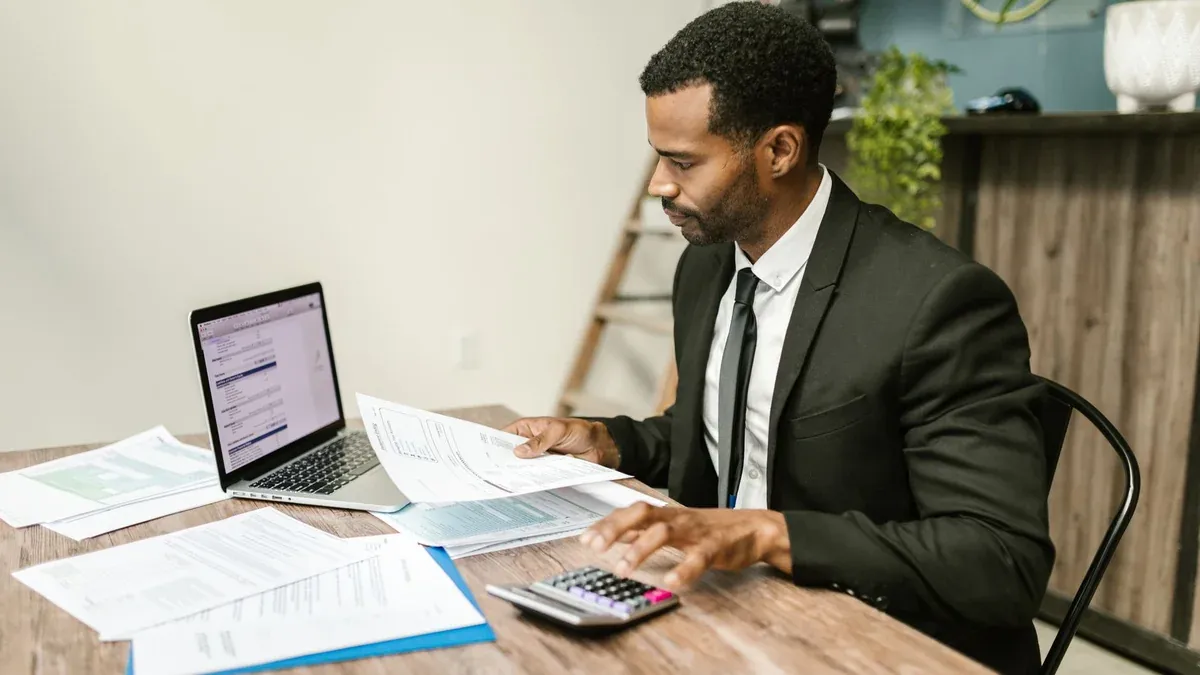 A businessman in a suit working on documents and using a laptop at an office desk.
