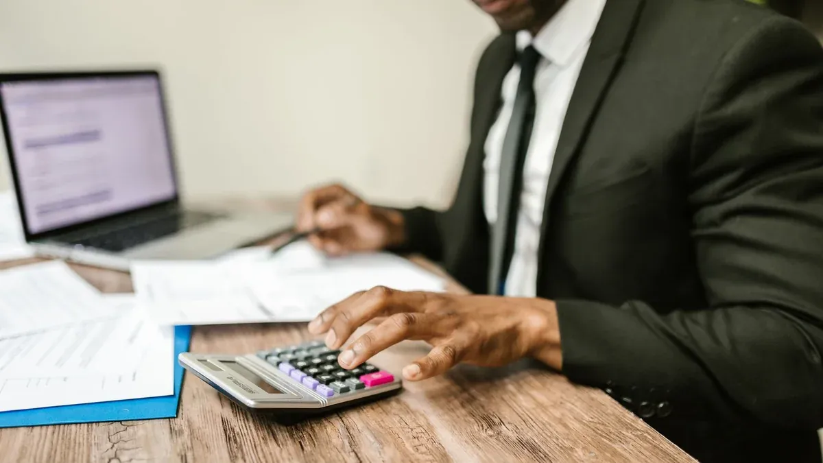 A businessman in a black suit using a calculator at a desk with financial documents and a laptop.