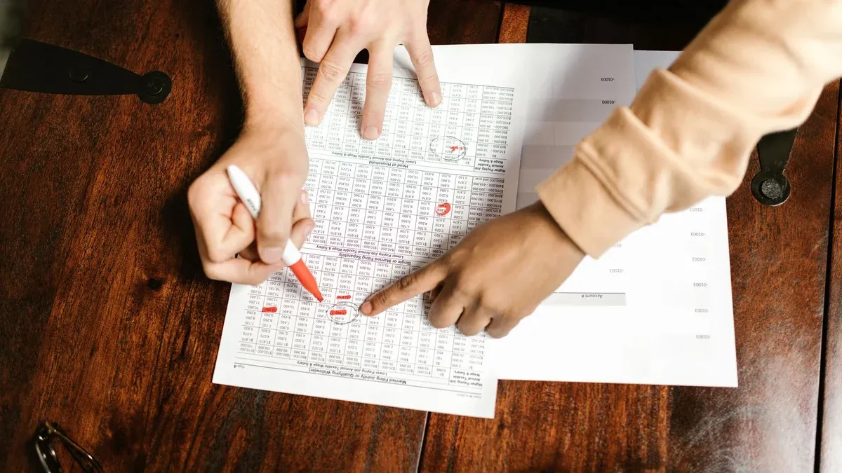 Two hands reviewing and marking financial documents with a red pen on a wooden table, top view.