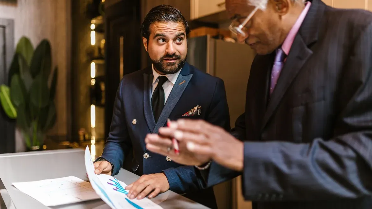 Two men in suits discuss financial documents with graphs indoors.