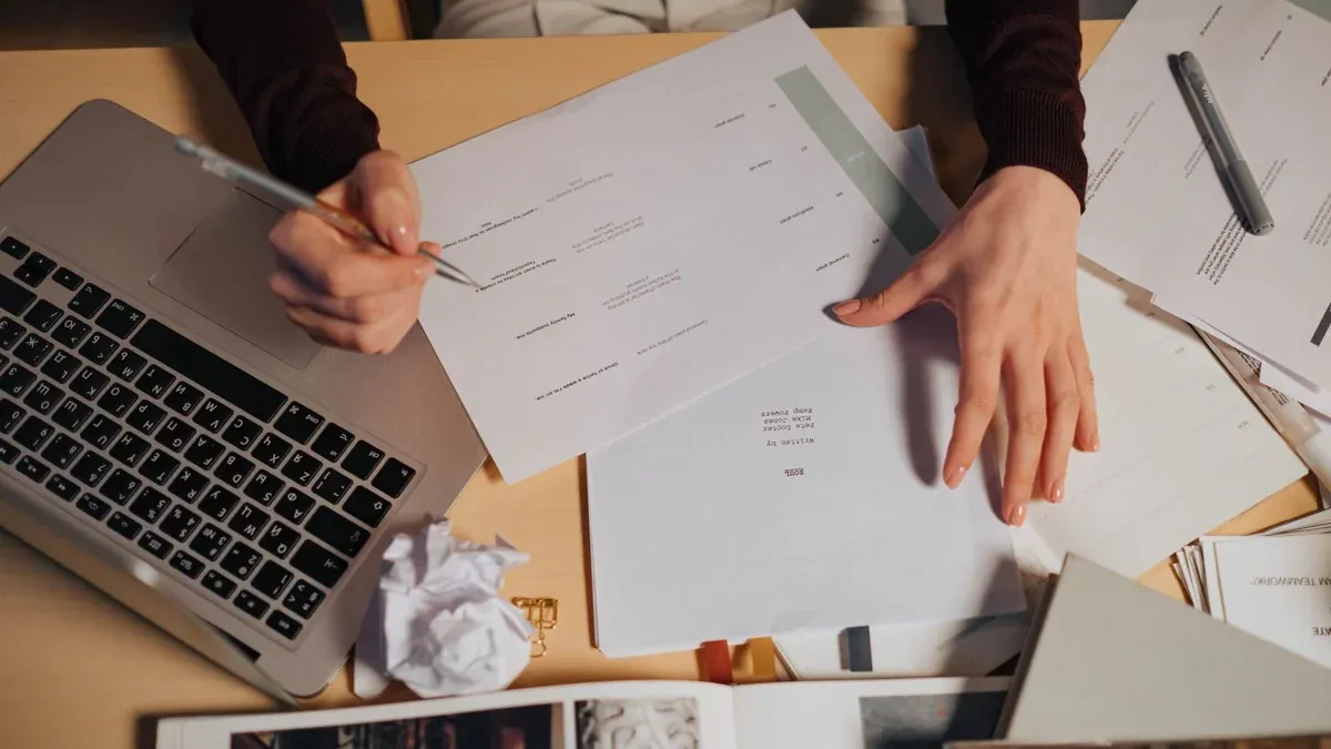 A person taking notes and working on a laptop surrounded by documents on a desk.