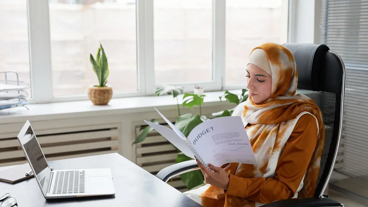 Muslim businesswoman in hijab working on budget documents at office desk.