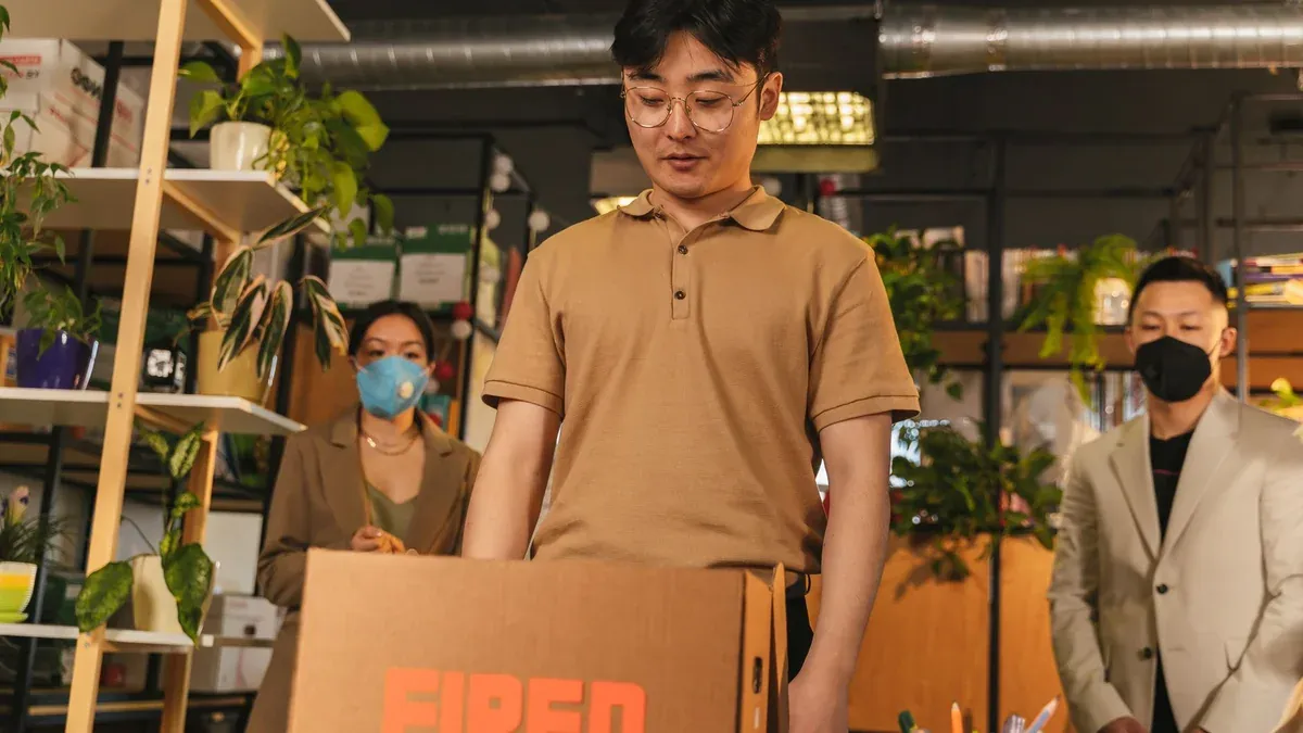 Man standing by cardboard box labeled 'fired', in an office surrounded by colleagues.