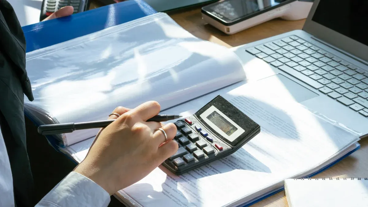 A person working on financial calculations using a calculator and laptop at an office desk.