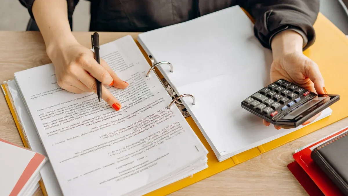 A woman reviewing documents and using a calculator on a wooden table.