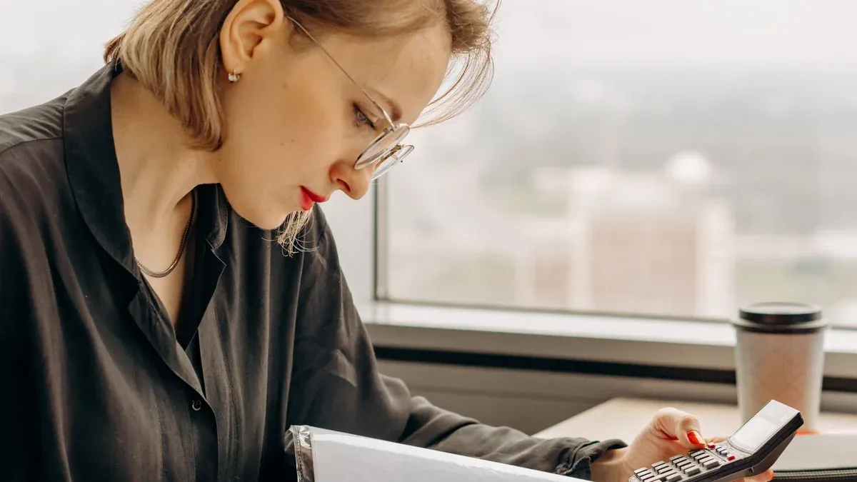 Businesswoman working with calculator and financial documents by window, focused and analytical.