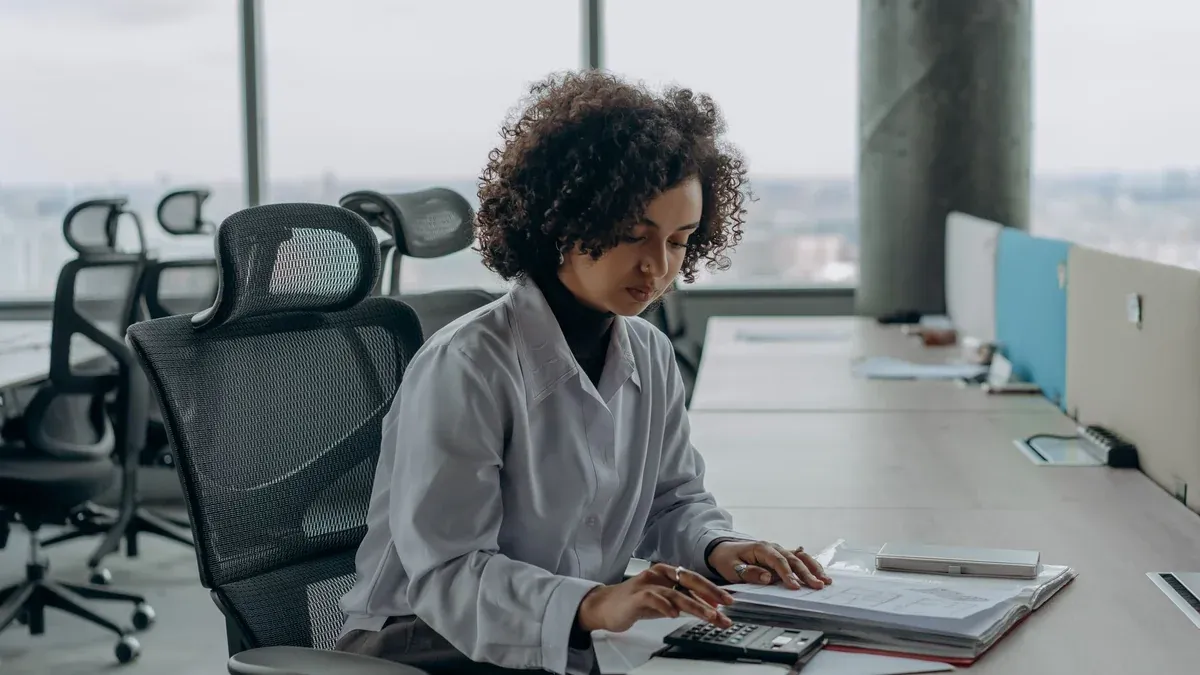A professional woman with curly hair calculating documents in a modern office setting.