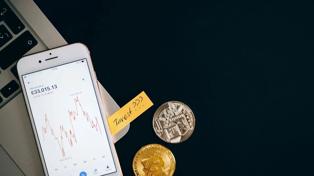 A smartphone displaying cryptocurrency prices beside coins and a laptop, symbolizing investment.