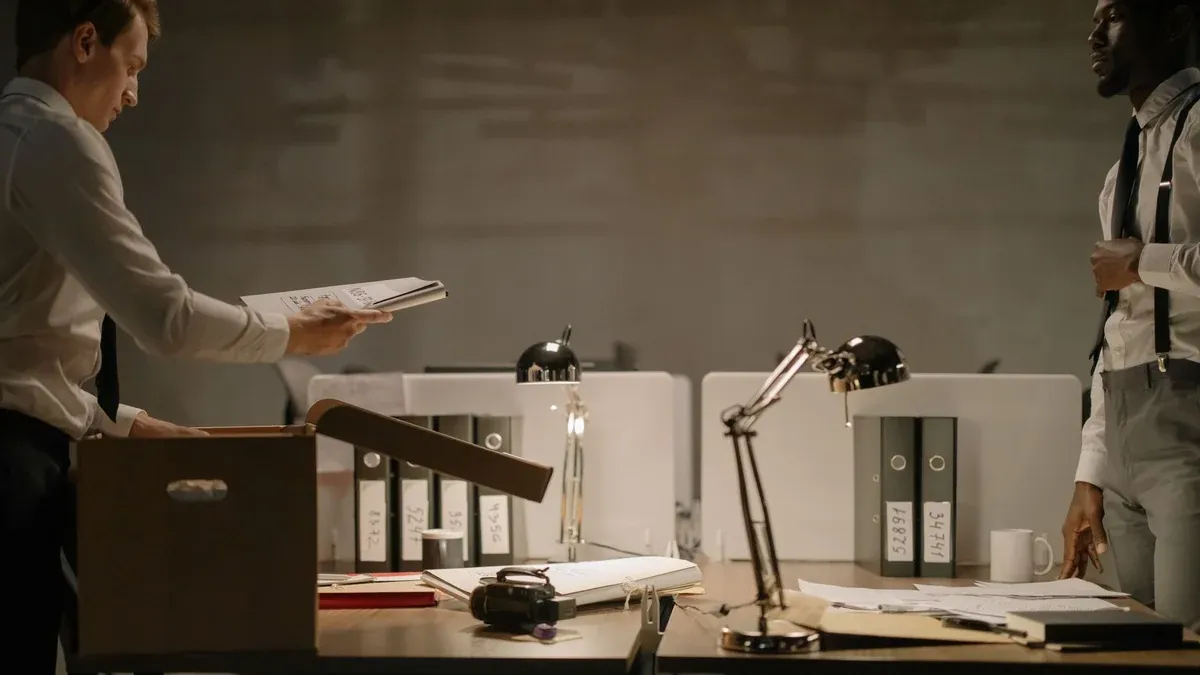 Two office workers sorting documents in a dimly lit workspace with several file binders.