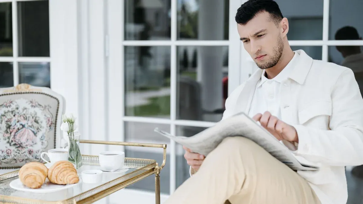 Elegant man reading a newspaper while enjoying breakfast with croissants and coffee on a patio.