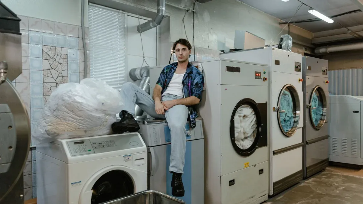 A man sits on a washing machine in a bustling laundry facility with industrial dryers.