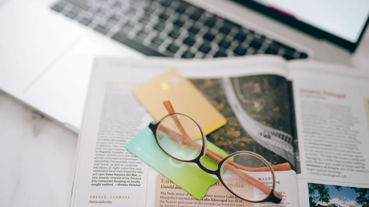 Overhead view of reading glasses, credit cards, and a magazine with a blurred laptop in the background.
