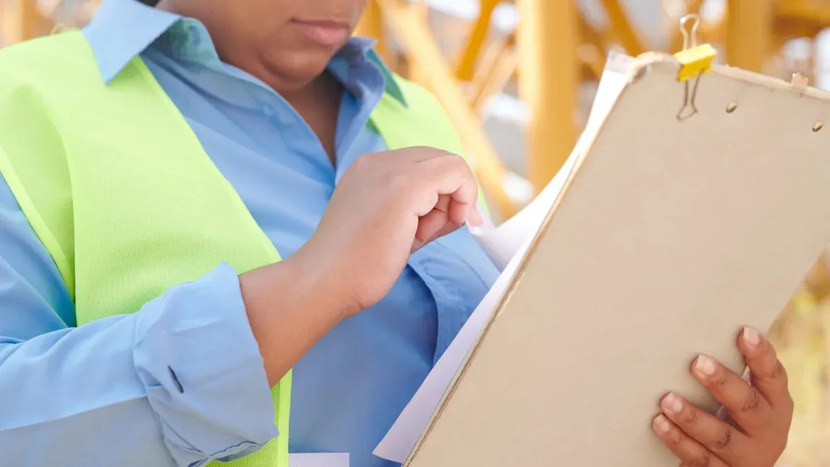 A female engineer in safety gear reviewing documents on a clipboard at a construction site.
