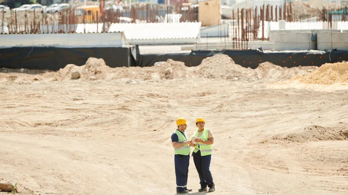 Two workers discussing plans on a sandy construction site, wearing safety gear.