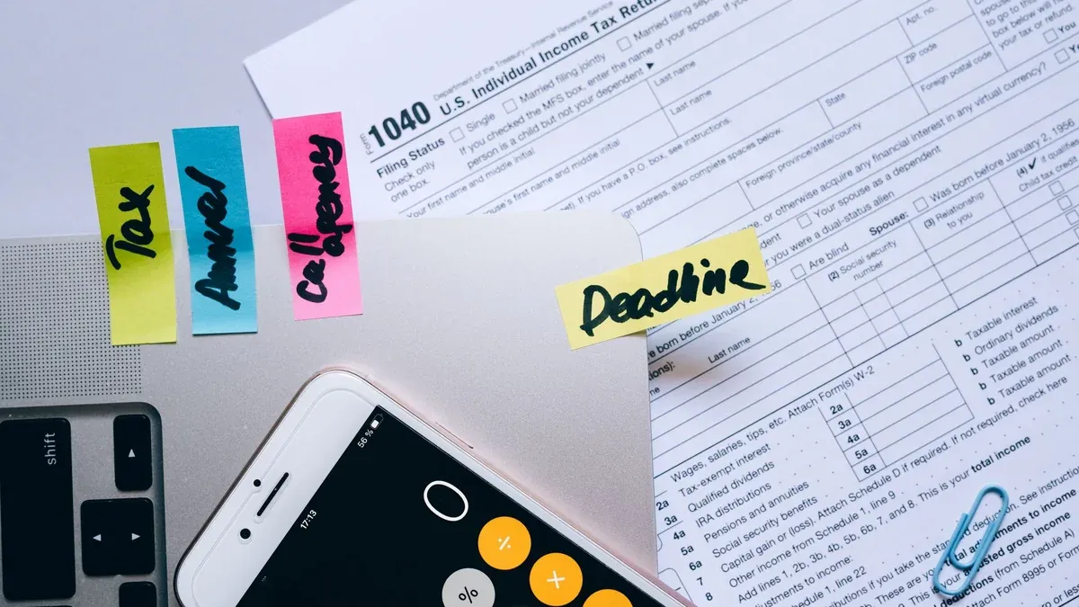 Office desk flat lay showing tax documents, calculator app on smartphone, sticky notes, and paperclips.